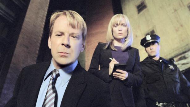 Zwei Detektive und ein Polizist stehen vor einem Gebäude.