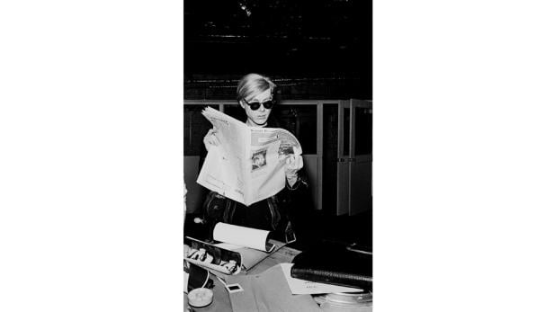 Andy Warhol liest eine Zeitung hinter einem Schreibtisch.