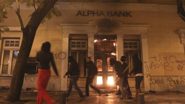 Vermummte Personen stehen vor einer brennenden Alpha Bank, die mit Graffiti beschmiert ist.