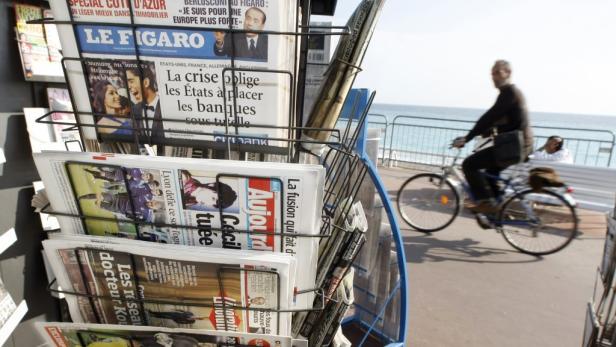 Ein Zeitungsstand mit verschiedenen französischen Zeitungen und ein Radfahrer im Hintergrund.