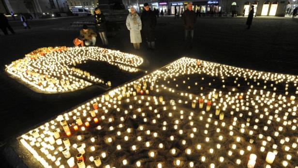 Viele Kerzen bilden ein Kreuz auf einem Platz, während Menschen im Hintergrund stehen.