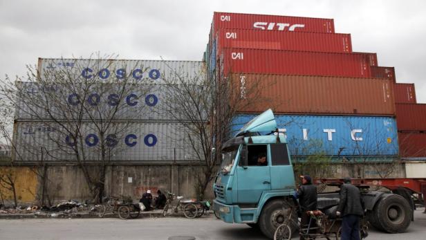 Ein blauer LKW steht vor einem Stapel von Frachtcontainern verschiedener Reedereien.