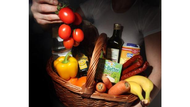 Ein Korb gefüllt mit Lebensmitteln wie Gemüse, Obst, Wurst und Ölflasche wird gehalten.