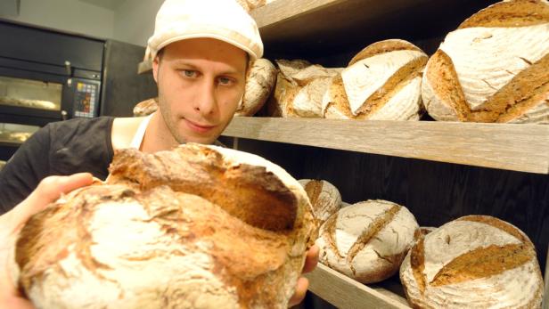Ein Bäcker präsentiert ein frisch gebackenes Brotlaib vor einem Regal voller Brote.