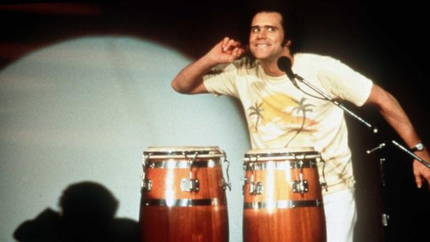 Jim Carrey spielt in einem weißen T-Shirt Congas auf einer Bühne.