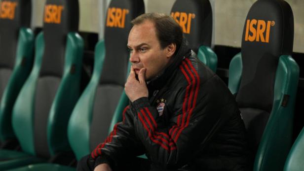 Ein Mann in einer Jacke des FC Bayern sitzt nachdenklich auf einer Bank.