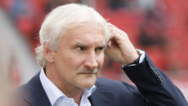 Ein Mann mit weißem Haar und Schnurrbart im Anzug kratzt sich am Kopf.