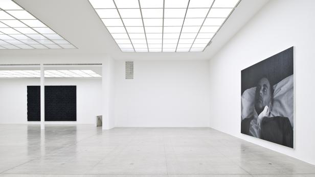 In einer Galerie hängen ein Porträt und zwei abstrakte schwarze Gemälde.