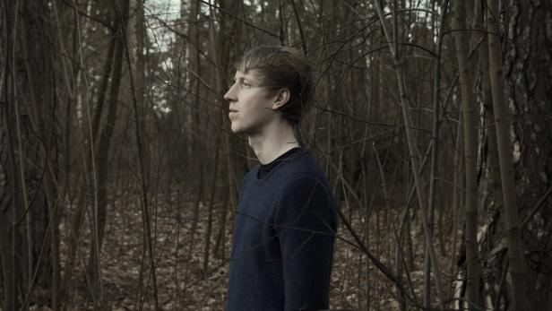 Ein junger Mann steht in einem dunklen Wald und blickt nach links.