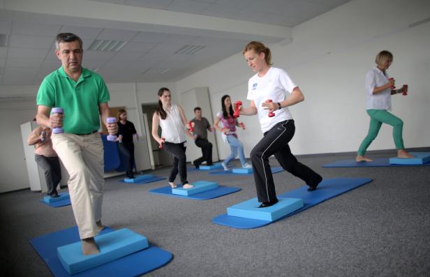 Eine Gruppe von Menschen trainiert mit Hanteln auf blauen Matten und Blöcken in einem hellen Raum.