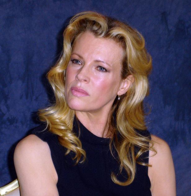 Kim Basinger mit blonden, gelockten Haaren vor einem blauen Hintergrund.
