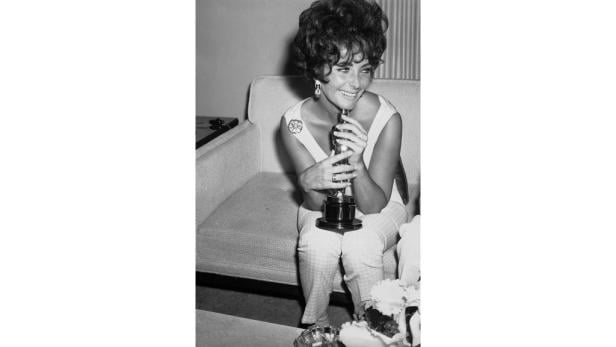 Elizabeth Taylor sitzt mit einem Oscar in den Händen auf einem Sofa.