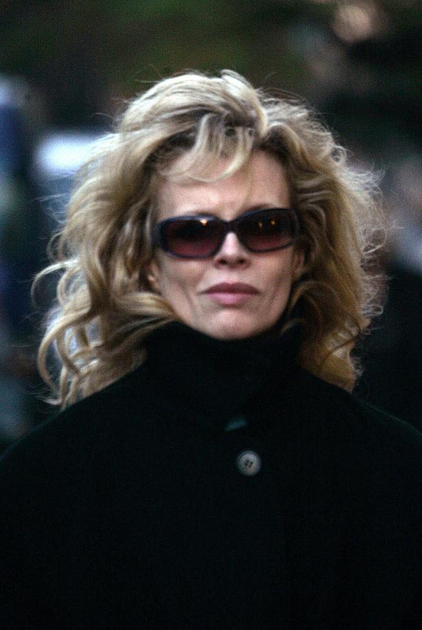 Eine Frau mit blonden, lockigen Haaren und Sonnenbrille trägt einen dunklen Mantel.