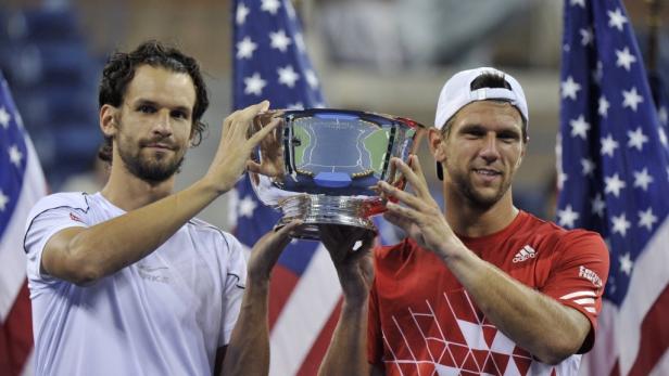 Zwei Tennisspieler halten eine silberne Trophäe vor US-amerikanischen Flaggen.