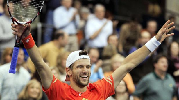 Ein lächelnder Tennisspieler mit erhobenen Armen und Tennisschläger feiert vor Publikum.