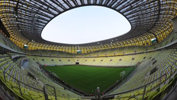 Blick in ein leeres Fußballstadion mit grüner Rasenfläche und gelben Sitzen.