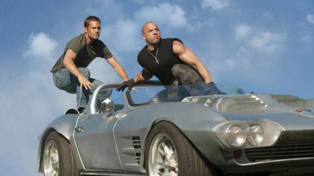 Vin Diesel und Paul Walker auf einem silbernen Sportwagen vor blauem Himmel.
