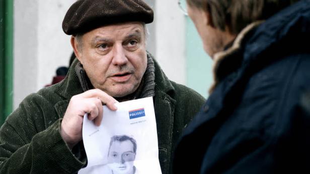 Ein Mann mit Cordjacke und Mütze zeigt einem anderen Mann ein Fahndungsfoto der Polizei.
