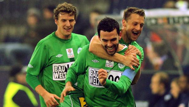 Drei Fußballspieler in grünen Trikots umarmen sich und feiern.