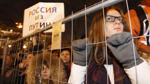 Eine Frau mit Brille blickt hinter einem Zaun auf eine Demonstration mit einem Schild „Russland ohne Putin“.