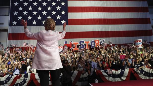 Hillary Clinton steht vor einer jubelnden Menge und einer riesigen US-Flagge.