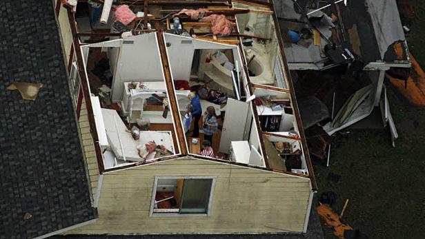 Ein Haus mit abgedecktem Dach und beschädigtem Innenraum wird von mehreren Personen begutachtet.