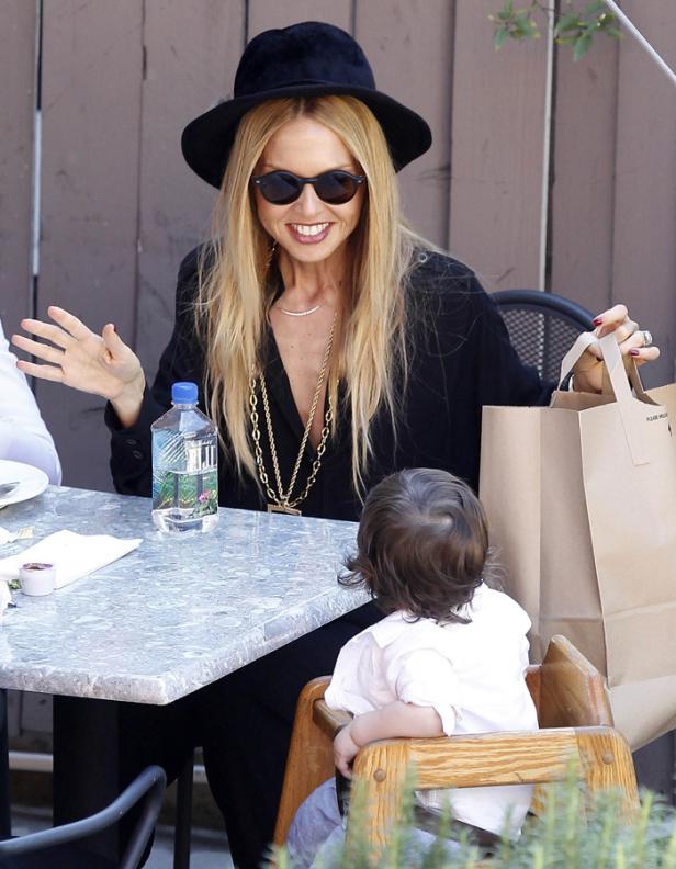 Eine Frau mit Hut und Sonnenbrille sitzt mit einem Kind an einem Tisch im Freien.
