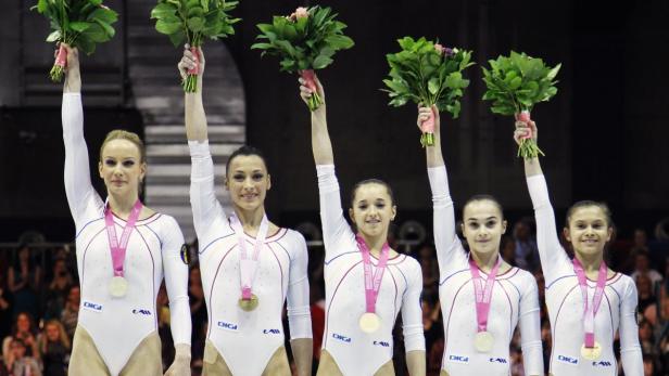 Fünf Turnerinnen mit Goldmedaillen und Blumensträußen feiern auf dem Podium.