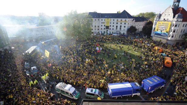 Eine große Menschenmenge in gelber Kleidung feiert auf einem Platz mit einem Banner „Der Meister kommt vom Borsigplatz!“.
