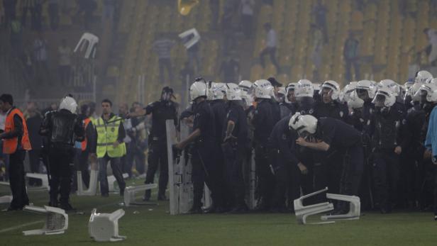 Polizisten in Schutzausrüstung stehen auf einem Fußballfeld, während Stühle durch die Luft fliegen.
