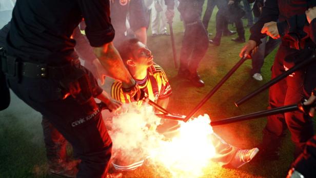 Polizisten umringen einen Mann mit einem Fußballtrikot, während eine Fackel brennt.