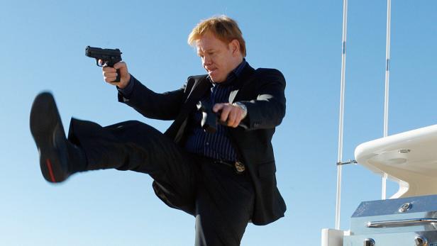 Ein Mann in einem Anzug zielt mit einer Pistole, während er ein Bein in die Luft wirft.