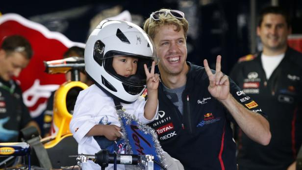 Sebastian Vettel mit einem Kind im Rennfahrerhelm, beide zeigen das Victory-Zeichen.