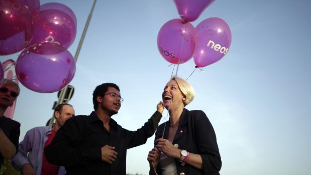 Eine lachende Frau hält lila „Neos“-Luftballons mit anderen Personen im Hintergrund.