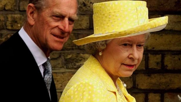 Königin Elisabeth II. mit gelbem Hut und Prinz Philip im Hintergrund.