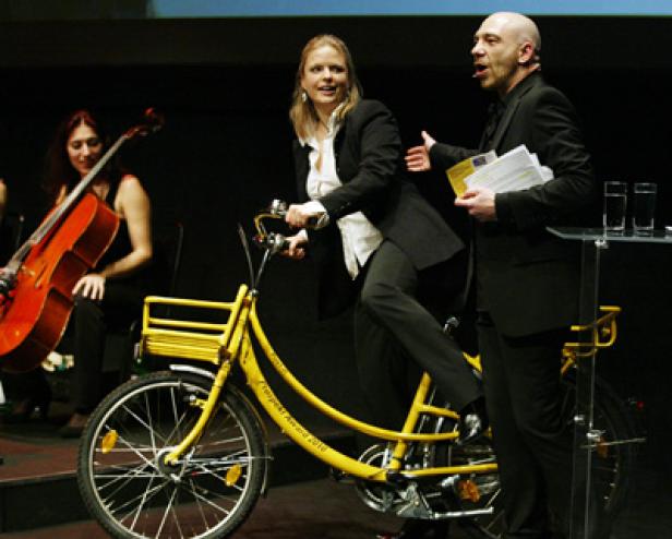 Eine Frau fährt auf einer Bühne auf einem gelben Fahrrad, während ein Mann eine Rede hält.