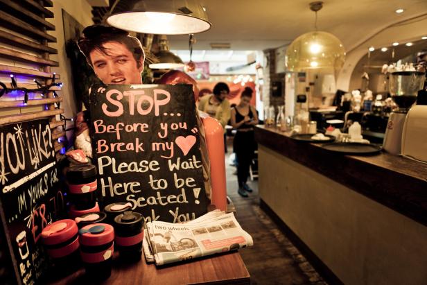 In einem Café steht eine Elvis-Pappfigur neben einer Tafel mit der Aufschrift „Stop… Before you break my heart. Please wait here to be seated!“.