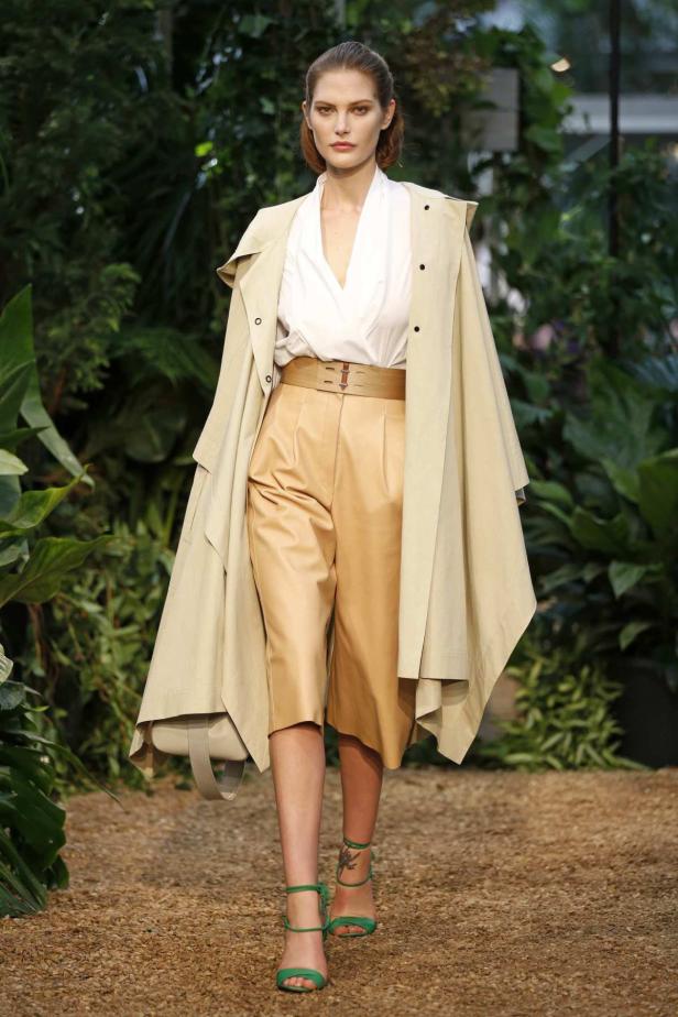 Eine Frau in beigefarbener Kleidung und grünen Sandalen läuft auf einem Laufsteg.