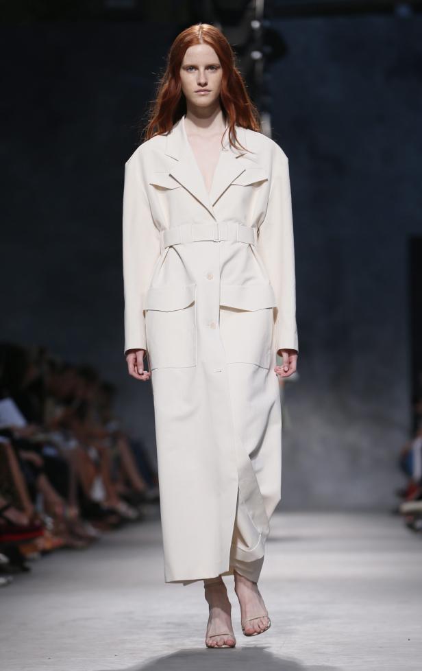 Eine rothaarige Frau präsentiert einen langen, hellen Mantel auf einem Laufsteg.