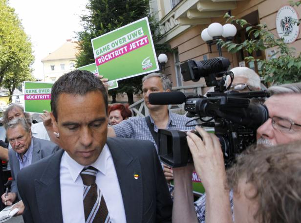 Ein Mann im Anzug wird von Demonstranten mit Schildern und Kameras umringt.