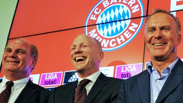 Drei Männer in Anzügen lächeln vor dem FC Bayern München Logo.