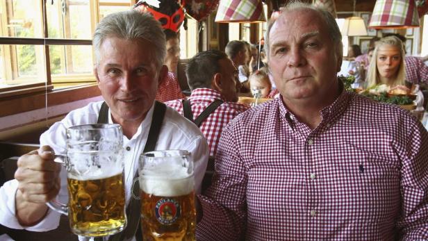 Zwei Männer in Tracht sitzen in einem Biergarten und halten Bierkrüge in die Kamera.