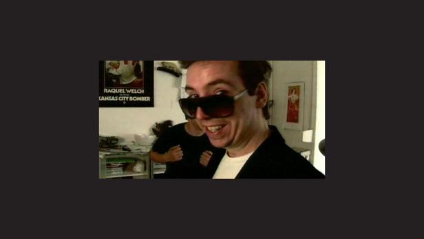 Ein Mann mit Sonnenbrille lächelt in die Kamera, im Hintergrund ein Poster von Raquel Welch.