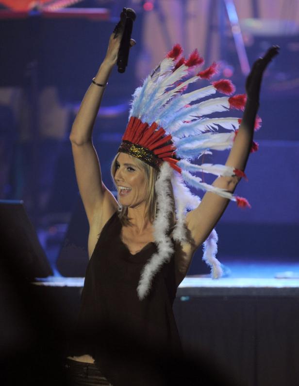 Heidi Klum mit einem indigenen Kopfschmuck und einem Mikrofon in der Hand auf einer Bühne.