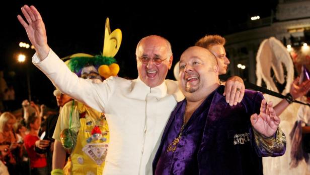Zwei Männer, der eine in Weiß, der andere in Lila, umarmen sich inmitten einer festlichen Menschenmenge.
