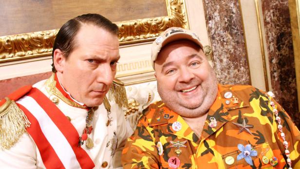 Zwei Männer sitzen nebeneinander, einer in einer Militäruniform, der andere in einem Hemd mit Camouflage-Muster und Ansteckern.