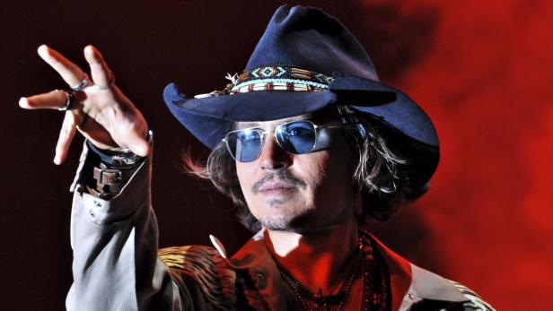 Johnny Depp mit Hut und Sonnenbrille gestikuliert vor rotem Hintergrund.