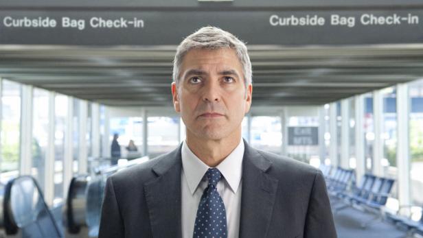 George Clooney in Anzug steht in einem Flughafenterminal.