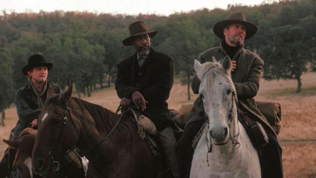 Drei Männer, darunter Morgan Freeman und Clint Eastwood, reiten auf Pferden durch eine hügelige Landschaft.