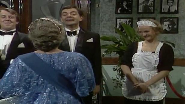 Rowan Atkinson und andere Schauspieler in einer Szene, in der eine Frau als Königin verkleidet ist.